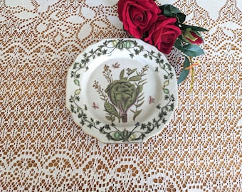 May include: A white ceramic plate with a green and brown floral design featuring a cabbage and other vegetables. The plate has a scalloped edge and is on a white lace tablecloth.