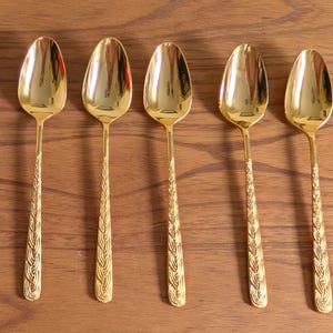 May include: Five gold-colored spoons with a reflective surface are arranged on a wooden surface. The spoon handles have a textured, woven design. The spoons are evenly spaced and appear to be made of metal.