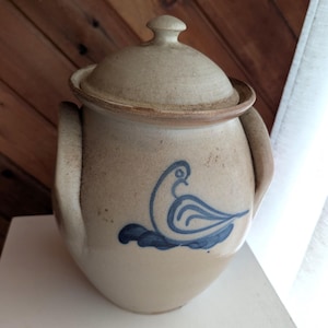 May include: A beige ceramic jar with a lid and two side handles. The jar features a blue bird design on the front. The jar is likely used for storage and is a decorative kitchen item.