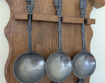 May include: Set of six pewter spoons with figures of people on the handles. The spoons are displayed on a wooden wall rack.
