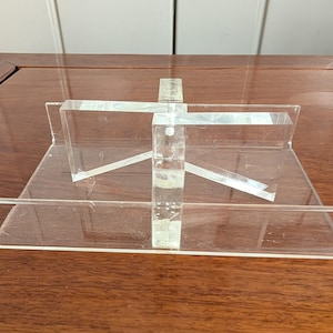 May include: Clear acrylic display stand with a rectangular base and a vertical support structure. Angled supports and a horizontal shelf create a tiered design. The stand is designed to hold items for display.