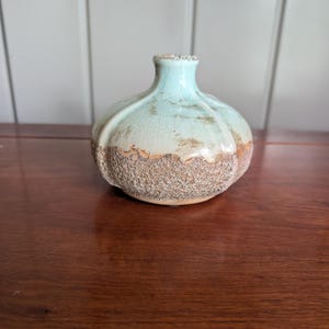 May include: A small, round ceramic vase with a pale mint green glazed upper section featuring a fine crackle pattern. The lower portion has a textured, rough, sandy brown finish. The vase has a narrow neck and a wide, bulbous body shape.