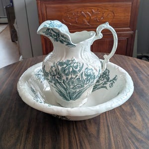 May include: Antique white porcelain pitcher and basin set. The pitcher has a curved spout and handle, and both pieces are decorated with a teal floral pattern. The basin has a scalloped edge. The set is displayed on a dark wooden table.