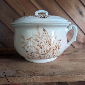 May include: Cream-colored antique chamber pot with a lid and handle. The pot features a brown floral design. The lid has a small handle. The pot is sitting on a wooden surface.