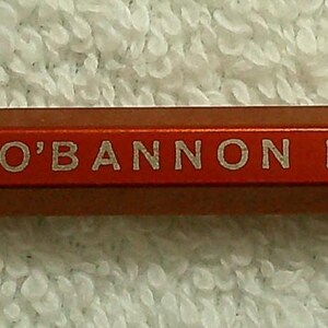 Może przedstawiać: Czerwony ołówek z białym napisem "USE O'BANNON PRODUCTS". Ołówek ma srebrny metalowy klips i srebrną metalową końcówkę.