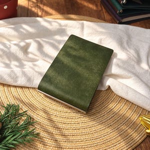 May include: A rectangular notebook with a dark olive green cover featuring a textured, matte finish. The notebook is closed and has a slim profile with a metallic edge visible at the bottom, presenting a clean and simple design for daily note-taking.