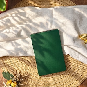 May include: A rectangular notebook with a solid dark green cover and a metallic binding edge. The notebook is positioned vertically, featuring a smooth, matte texture on the front surface, capturing light and shadow patterns across its surface.