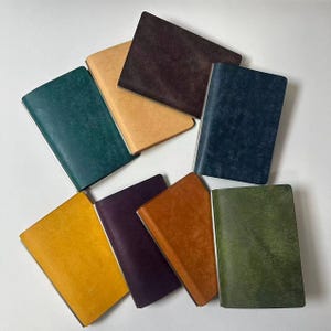 Puede incluir: Una colección de ocho cuadernos rectangulares con cubiertas texturizadas en varios colores, incluyendo verde azulado, bronceado, marrón oscuro, azul marino, amarillo, morado, naranja y verde oliva, dispuestos en un patrón superpuesto.