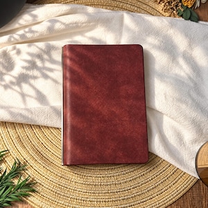 Könnte beinhalten: Ein rechteckiges Notizbuch mit einem tiefroten, strukturierten Einband und einem metallischen Buchrücken. Der vordere Einband hat eine glatte, matte Oberfläche mit subtilen Farbnuancen und bietet ein schlichtes Design zum Schreiben oder Notieren.