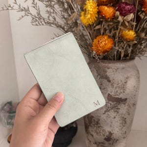 May include: A rectangular, light sage green compact mirror with a textured, matte finish. The bottom right corner features a small, embossed capital letter M in a serif font. The mirror has a slim, hinged design with a metallic silver edge.