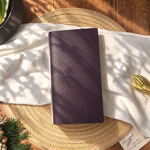 May include: A rectangular, dark purple notebook with a smooth, matte cover. The notebook is positioned vertically, featuring a clean, solid-colored front surface with sharp, defined edges and a slim profile, presenting a simple and functional design.