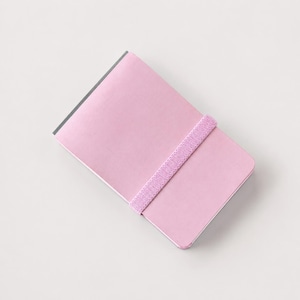 May include: A rectangular notebook with a solid light pink cover and rounded corners. The notebook is secured by a matching light pink elastic band that wraps horizontally around the center of the cover, keeping the pages closed and protected.