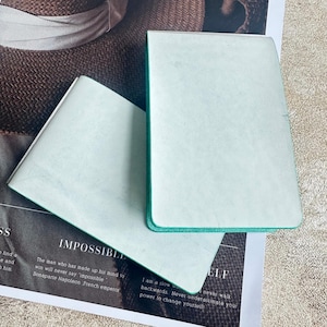 Può includere: Due quaderni verde chiaro con bordi turchese sono impilati su una rivista. Il quaderno superiore è aperto, rivelando pagine bianche. La rivista ha il testo "IMPOSSIBLE" e una citazione di Napoleone.