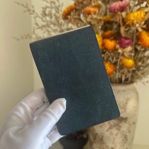 May include: A rectangular, dark charcoal-coloured object with a matte, slightly textured surface. The item has clean, straight edges and a flat, solid appearance, held in a white-gloved hand, presenting a simple and uniform dark grey finish.