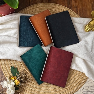May include: Five rectangular notebooks with textured covers in dark blue, tan, black, teal, and deep red. Each notebook features a clean white spine edge and a simple, solid-colored front cover design, arranged in a layered, overlapping fan-like formation.