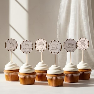 Könnte beinhalten: Sechs Cupcakes mit weißem Zuckerguss und dekorativen Tortenauflegern. Die runden Aufleger haben braune und weiße gestreifte Ränder und zeigen Illustrationen und Text, darunter "a baby is brewing" und "Olivia's baby shower".
