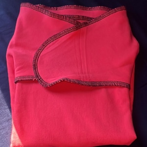 Può includere: Una copertina per neonati in tessuto rosa acceso con cuciture nere sui bordi. La copertina è piegata a metà e si trova su una superficie blu scuro.