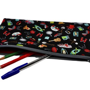 May include: Black pencil case with a camping-themed pattern, featuring illustrations of a guitar, campfire, and kayak. The case is open, revealing a red pencil, a green pencil, and a blue pen.