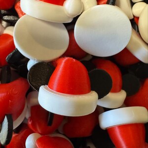 May include: A collection of small, 3D-printed ornaments shaped like red Santa hats with white brims and black circular ears. Each piece features a smooth, flat white circular base on the top, creating a uniform appearance across the pile of items.