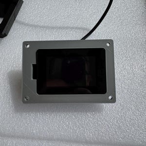 May include: A rectangular, gray electronic component with a black screen. The component has four mounting holes and a small cutout on one side. Black wires extend from the top. The item is on a white, textured surface.