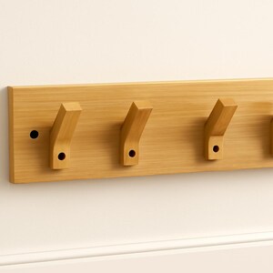 Puede incluir: Un perchero de pared de madera con cuatro ganchos angulados. El perchero es de madera de color claro y tiene forma rectangular. Cada gancho tiene un pequeño agujero redondo. Está montado en una pared blanca.