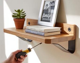 Puede incluir: Estante flotante de madera con una pequeña suculenta en maceta, dos libros y una foto enmarcada con el texto "family memories". El estante se está instalando con un destornillador. El estante tiene soportes de metal negro.