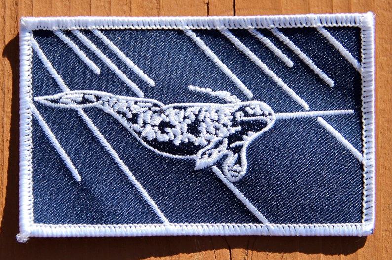 Peut inclure: &Eacute;cusson brod&eacute; repr&eacute;sentant un narval avec une longue d&eacute;fense, sur un fond bleu fonc&eacute; avec des lignes diagonales blanches. Le narval est d&eacute;taill&eacute; avec des coutures blanches, et l'&eacute;cusson a une bordure blanche.