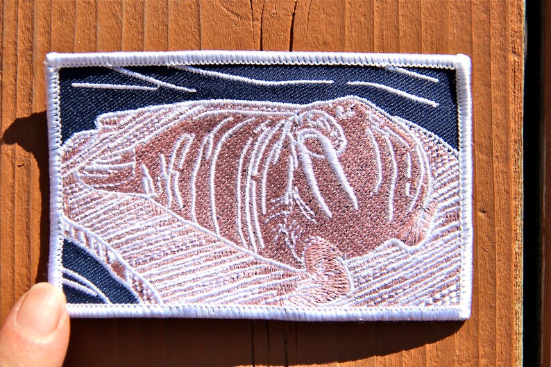 May include: White embroidered patch of a walrus on a dark blue background. The walrus is lying down with its tusks visible.