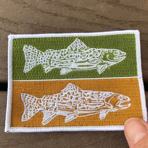 Pode incluir: Um remendo bordado branco com duas trutas, uma em um fundo verde e outra em um fundo dourado. As trutas s&atilde;o estilizadas e t&ecirc;m um design detalhado.