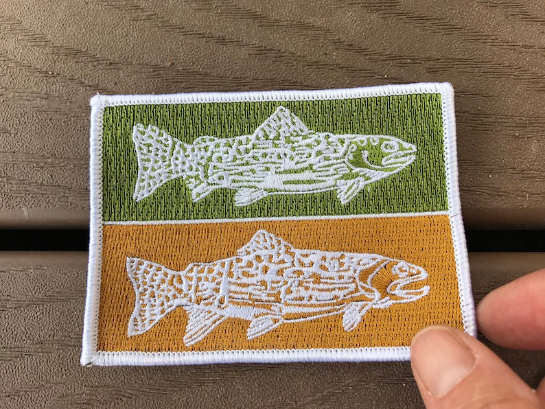 Pode incluir: Um remendo bordado branco com duas trutas, uma em um fundo verde e outra em um fundo dourado. As trutas s&atilde;o estilizadas e t&ecirc;m um design detalhado.