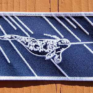 Peut inclure: &Eacute;cusson brod&eacute; repr&eacute;sentant un narval avec une longue d&eacute;fense, sur fond bleu marine avec des lignes diagonales blanches. L'&eacute;cusson a une bordure blanche et est de forme rectangulaire.