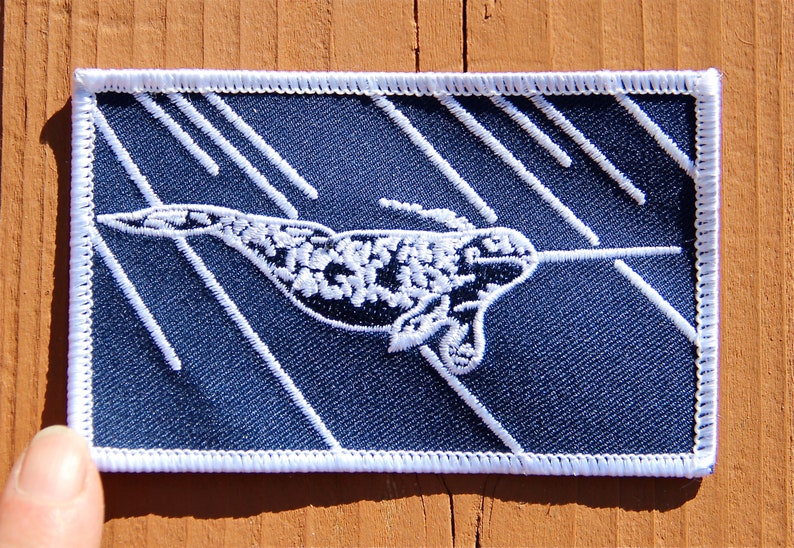 Peut inclure: &Eacute;cusson brod&eacute; repr&eacute;sentant un narval avec une longue d&eacute;fense, sur fond bleu marine avec des lignes diagonales blanches. L'&eacute;cusson a une bordure blanche et est de forme rectangulaire.