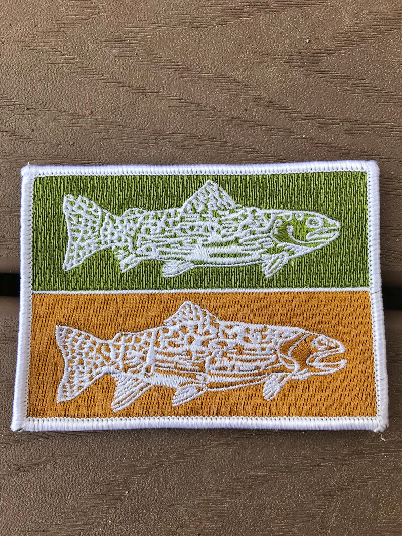 Pode incluir: Um remendo bordado branco com dois peixes, um em um fundo verde e outro em um fundo dourado. Os peixes s&atilde;o estilizados e t&ecirc;m um padr&atilde;o detalhado.