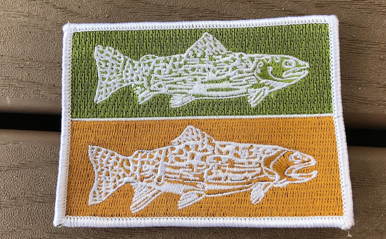 Pode incluir: Um remendo bordado branco com duas trutas, uma em um fundo verde e a outra em um fundo dourado.