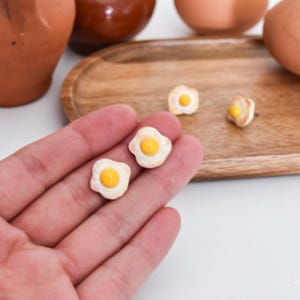Op de afbeelding: Miniatuur gebakken ei oorbellen met een gele dooier en wit eiwit. De oorbellen worden in een hand gehouden, met andere keramische items op de achtergrond, waaronder een houten dienblad en terracotta potten.