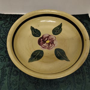 May include: A large, cream-colored ceramic bowl with a brown rim and a hand-painted design of a purple flower with green leaves.