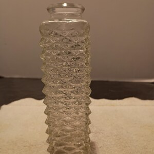 May include: A clear glass bottle with a textured, diamond-patterned exterior. The bottle has a cylindrical shape with a slightly flared base and a small, flat lid. The glass appears to be vintage.