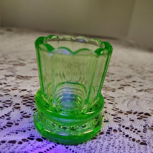May include: A small, green glass vase with a scalloped rim and a fluted design. The vase has a base with two rings of decorative detail.