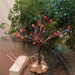 May include: A decorative tree sculpture with dark brown textured branches adorned with small, translucent, multi-colored spherical beads in shades of red, blue, green, and orange. The tree stands on a tan base and includes a white battery pack.