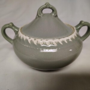 May include: A green ceramic sugar bowl with a white lid and a white decorative design around the rim. The bowl has two handles.