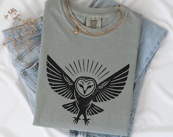 May include: A sage green t-shirt with a black owl graphic, featuring outstretched wings and radiating lines. The shirt is folded over light blue denim jeans. A gold necklace and dried flowers are also visible.