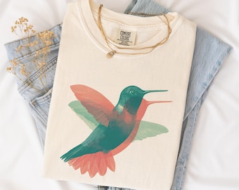 May include: Cream-colored t-shirt with a vibrant hummingbird design in shades of teal, coral, and green. The shirt is paired with light-wash denim jeans and a delicate gold necklace. The Comfort Colors tag is visible.