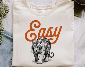 May include: A cream-colored t-shirt featuring the word Easy in a bold, orange, cursive font positioned above a detailed, black and white illustration of a tiger walking forward. The design is centered on the front of the garment.
