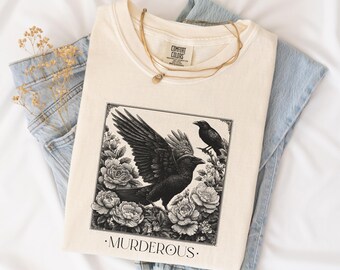 May include: A cream-colored t-shirt featuring a black and white graphic of two crows surrounded by roses within a square frame. The word MURDEROUS is printed in black capital letters below the frame. A gold chain necklace rests on the neckline.