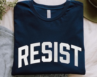 May include: A dark navy blue crew neck t-shirt featuring the word RESIST printed in large, white, capital letters in an arched layout across the chest. The shirt has a standard short-sleeve design and a visible brand label inside the neckline.