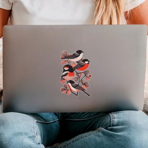 A beautiful and minimal illustration of four finches perched in a tree. Design is black, red and cream, printed on a kiss-cut vinyl sticker with a transparent  border. Sticker is shown on the back of an open silver laptop as a young lady is working.