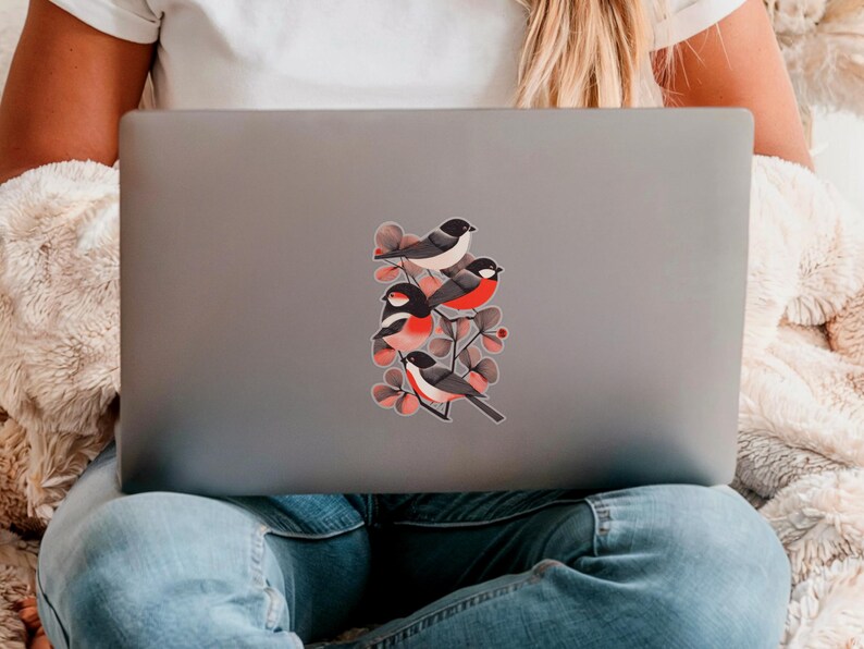 A beautiful and minimal illustration of four finches perched in a tree. Design is black, red and cream, printed on a kiss-cut vinyl sticker with a transparent  border. Sticker is shown on the back of an open silver laptop as a young lady is working.