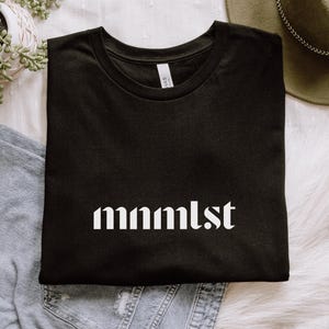 A minimalist typographic design that reads "mnmlst". Design is white on a Black Bella+Canvas 3001 t-shirt. Garment is folded with jeans, fedora and succulent plant on a white background.