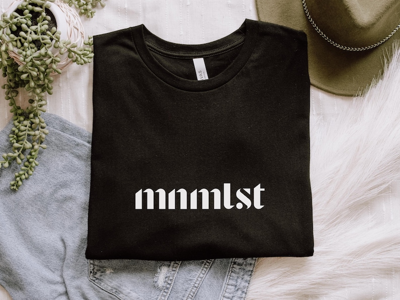 A minimalist typographic design that reads "mnmlst". Design is white on a Black Bella+Canvas 3001 t-shirt. Garment is folded with jeans, fedora and succulent plant on a white background.