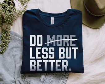 May include: Navy blue t-shirt with the text "DO MORE LESS BUT BETTER" in white and grey block letters. The shirt is folded on top of a pair of blue jeans and a grey sweatshirt. A green hat and plant are in the background.